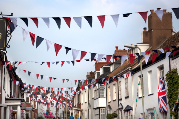 A street with quaint cottages running on either side with red, white and blue bunting strung between the cottage roofs.