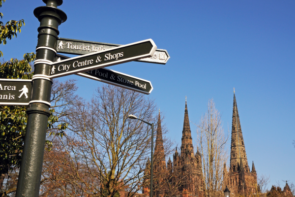 A sign pointing to the city centre shops, Stowe pool and tourist information, with Lichfield cathedral in the background against a blue sky