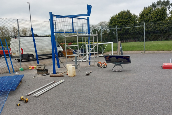 A photograph of the Multi Use Games Area at Burntwood Leisure Centre. It is a grey court outside, surrounded by greenery. In frame blue posts are being erected, with construction equipment nearby and a white van in the background.