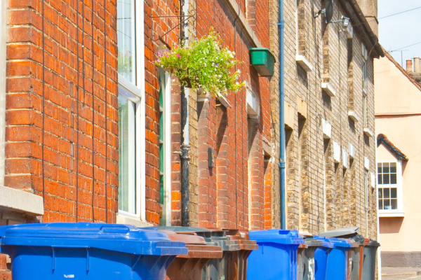 A street of brown brick houses with hanging baskets and a row of blue, brown and black bins lined up in front of the houses.