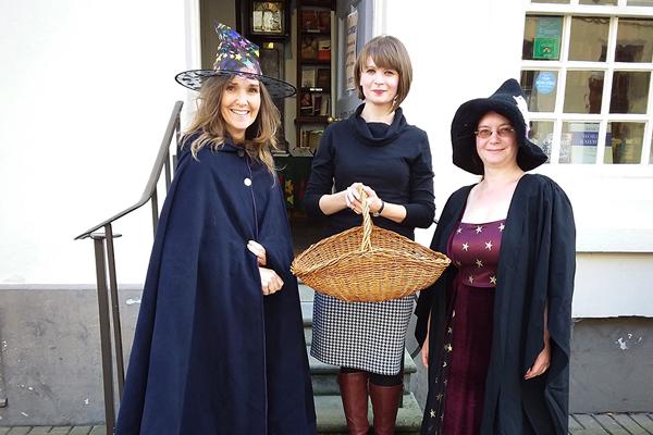 Two women in cloaks and witches hat either side of a woman holding a basket.